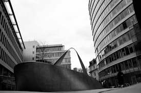 A modern urban landscape featuring a large, abstract metal sculpture positioned between two contemporary high-rise buildings. The sculpture curves upward with sleek lines, contrasting against the glass and steel architecture surrounding it. A person is walking nearby, adding a sense of scale to the scene. The overcast sky creates a stark black and white visual.