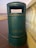 A green cylindrical outdoor trash can stands on a brown tiled surface, adjacent to a white balustrade. The can has a rectangular opening at the top for waste disposal and is set against a backdrop featuring a soft gradient sky.