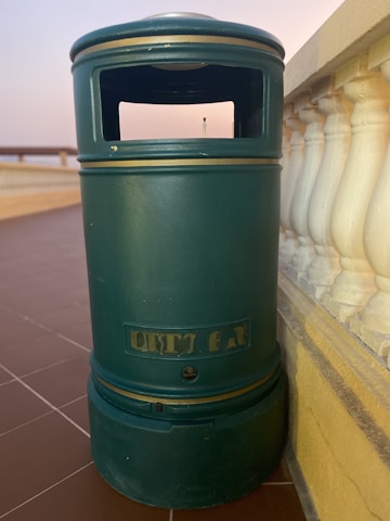 A green cylindrical outdoor trash can stands on a brown tiled surface, adjacent to a white balustrade. The can has a rectangular opening at the top for waste disposal and is set against a backdrop featuring a soft gradient sky.