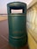 A green cylindrical outdoor trash can stands on a brown tiled surface, adjacent to a white balustrade. The can has a rectangular opening at the top for waste disposal and is set against a backdrop featuring a soft gradient sky.