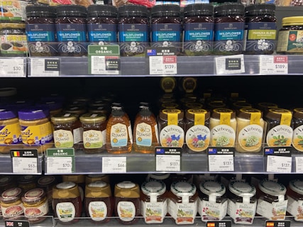 Shelves stocked with various brands of honey jars and bottles, including products labeled as organic and manuka blend from different countries such as New Zealand and Belgium. The display includes a range of container types, including glass jars and plastic squeeze bottles.