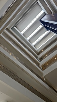 The image captures the interior view of a multi-story building, showcasing an architectural perspective looking up. The shot reveals multiple levels with a series of balconies and railings lining the open central space. Natural light enters through rectangular skylights positioned near the ceiling.