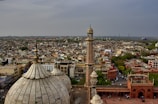 An aerial view of a densely populated city with a prominent mosque featuring large domed roofs and a tall minaret. The cityscape includes a mixture of residential and commercial buildings, with lush green areas scattered throughout. The sky is overcast, creating a muted atmosphere.