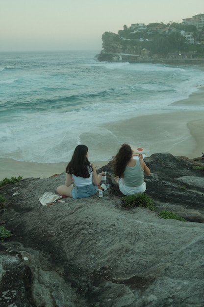 two women sitting on a rock overlooking the ocean