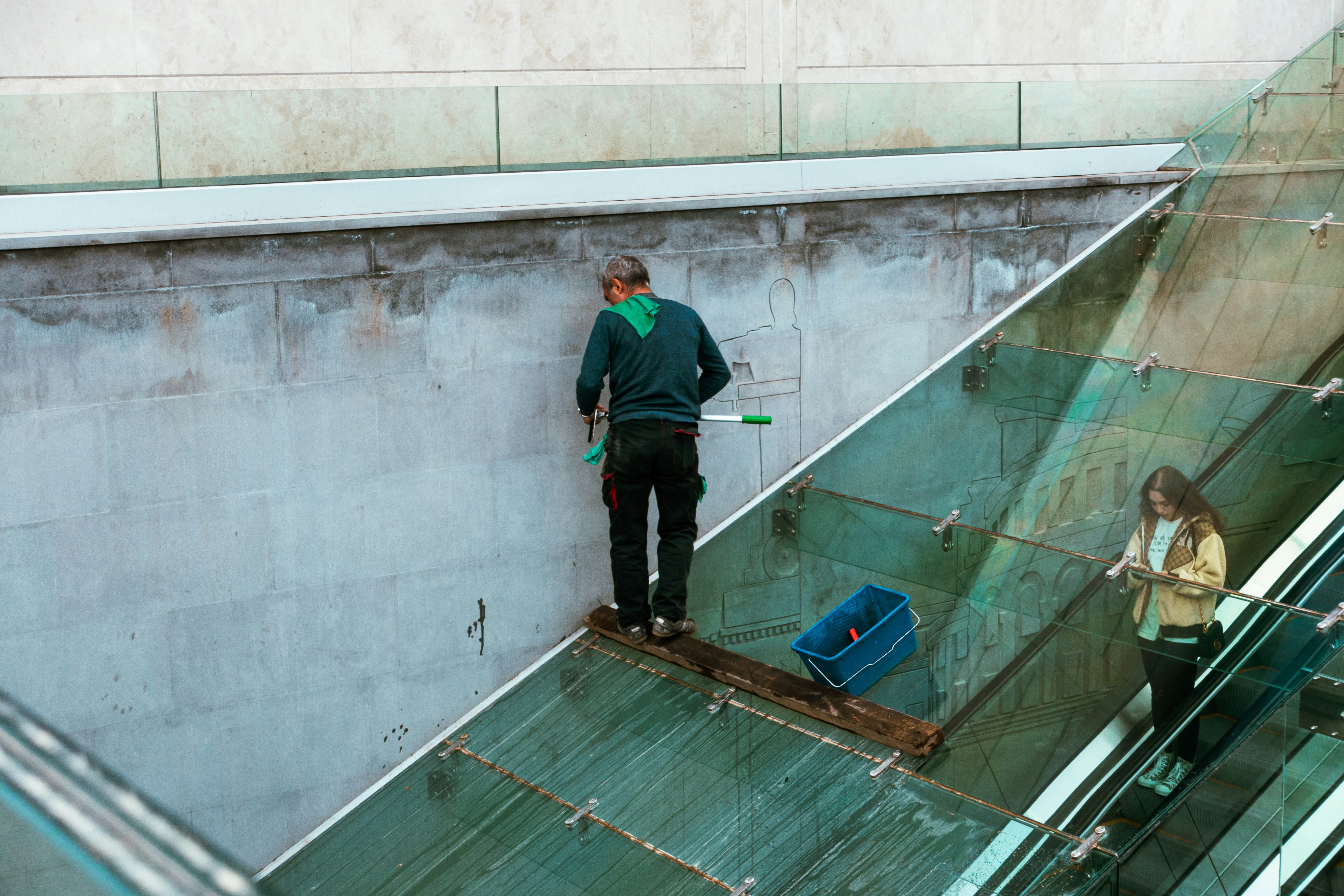 A person is cleaning a glass surface on a modern building using a squeegee. They stand on a narrow ledge with a safety harness attached. Another person is on an escalator inside the building, partially visible through the glass.