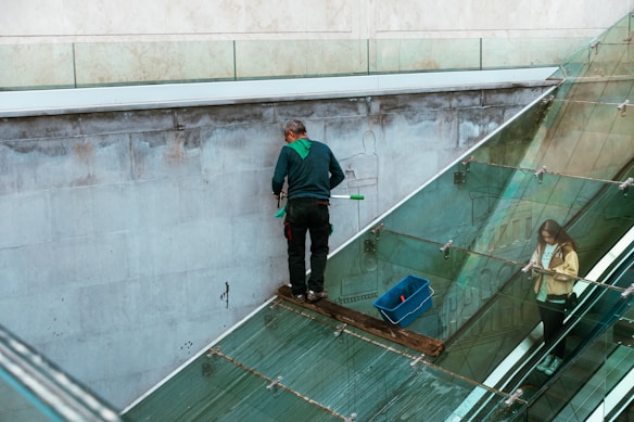 A person is cleaning a glass surface on a modern building using a squeegee. They stand on a narrow ledge with a safety harness attached. Another person is on an escalator inside the building, partially visible through the glass.