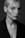 A black and white portrait of a person with light-colored hair partially obscuring one eye. The shot focuses on the face, highlighting strong cheekbones and a neutral expression. The individual is wearing a dark coat, and the overall lighting creates a soft contrast on the skin.
