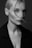 A black and white portrait of a person with light-colored hair partially obscuring one eye. The shot focuses on the face, highlighting strong cheekbones and a neutral expression. The individual is wearing a dark coat, and the overall lighting creates a soft contrast on the skin.