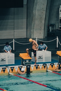 A swimmer wearing a yellow cap is standing on the starting block, preparing for a swim race in an indoor pool. Two officials are standing nearby at the edge of the pool, overseeing the event. The pool is marked with lane dividers and there are two other swimmers visible in the water near the edge.