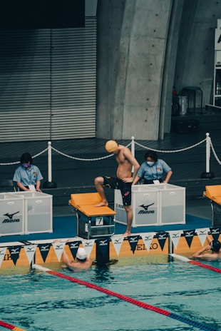 A swimmer wearing a yellow cap is standing on the starting block, preparing for a swim race in an indoor pool. Two officials are standing nearby at the edge of the pool, overseeing the event. The pool is marked with lane dividers and there are two other swimmers visible in the water near the edge.