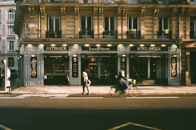 A street scene in front of a charming bakery and patisserie with classic Parisian architecture. A person walks on the sidewalk while another rides past on a bicycle. The facade of the building features a large window display and signage reading 'Pâtissier' and 'Boulanger', indicating a focus on pastry and bakery items.