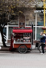 A mobile coffee cart with a red canopy is parked on a street. An assortment of coffee cups is displayed, and a menu is visible. A tree with bare branches overhangs the cart. A person wearing a blue and white jacket and dark pants is standing nearby, seemingly using a phone.