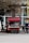 A mobile coffee cart with a red canopy is parked on a street. An assortment of coffee cups is displayed, and a menu is visible. A tree with bare branches overhangs the cart. A person wearing a blue and white jacket and dark pants is standing nearby, seemingly using a phone.