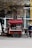 A mobile coffee cart with a red canopy is parked on a street. An assortment of coffee cups is displayed, and a menu is visible. A tree with bare branches overhangs the cart. A person wearing a blue and white jacket and dark pants is standing nearby, seemingly using a phone.