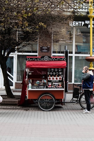 A mobile coffee cart with a red canopy is parked on a street. An assortment of coffee cups is displayed, and a menu is visible. A tree with bare branches overhangs the cart. A person wearing a blue and white jacket and dark pants is standing nearby, seemingly using a phone.