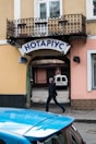 A street scene featuring a notary's office sign in Cyrillic letters mounted above an archway leading to a courtyard. A man is walking past under the arch, and a blue car is parked nearby. The building has a wrought iron balcony and pastel-colored walls.
