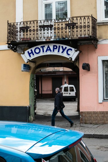 A street scene featuring a notary's office sign in Cyrillic letters mounted above an archway leading to a courtyard. A man is walking past under the arch, and a blue car is parked nearby. The building has a wrought iron balcony and pastel-colored walls.
