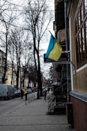A street view with bare trees lining the sidewalk. A Ukrainian flag is prominently displayed on a building to the right. Several pedestrians walk along the paved sidewalk, and cars are parked on the side of the road. The buildings are a mix of old architectural styles.