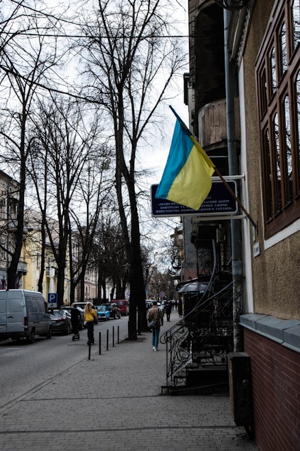 A street view with bare trees lining the sidewalk. A Ukrainian flag is prominently displayed on a building to the right. Several pedestrians walk along the paved sidewalk, and cars are parked on the side of the road. The buildings are a mix of old architectural styles.