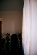 A dimly lit room with a clothing rack holding various garments, including shirts and jackets. A large white curtain covers part of the window, allowing some light to filter into the room.