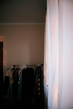 A dimly lit room with a clothing rack holding various garments, including shirts and jackets. A large white curtain covers part of the window, allowing some light to filter into the room.