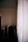 A dimly lit room with a clothing rack holding various garments, including shirts and jackets. A large white curtain covers part of the window, allowing some light to filter into the room.