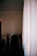 A dimly lit room with a clothing rack holding various garments, including shirts and jackets. A large white curtain covers part of the window, allowing some light to filter into the room.