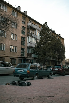 A residential street with an older apartment building featuring balconies. Several parked cars are visible along the sidewalk, and there are tall trees near the building. The building facade is beige with multiple windows, and there is a reddish-brown pavement in the foreground with some construction materials.