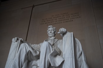 A large, seated marble statue of a man with a beard, wearing a suit and sitting on a high-backed chair. The surrounding structure is imposing with a high ceiling and inscription on the wall behind the statue.