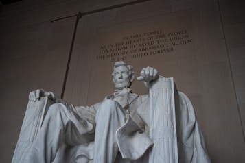A large, seated marble statue of a man with a beard, wearing a suit and sitting on a high-backed chair. The surrounding structure is imposing with a high ceiling and inscription on the wall behind the statue.