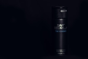 A sleek black cylindrical deodorant canister displaying the text 'David Beckham - The Essence'. The can has a glossy texture, with minimalist design elements, set against a dark background.