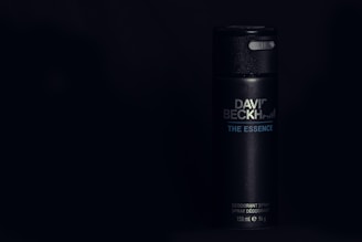 A sleek black cylindrical deodorant canister displaying the text 'David Beckham - The Essence'. The can has a glossy texture, with minimalist design elements, set against a dark background.