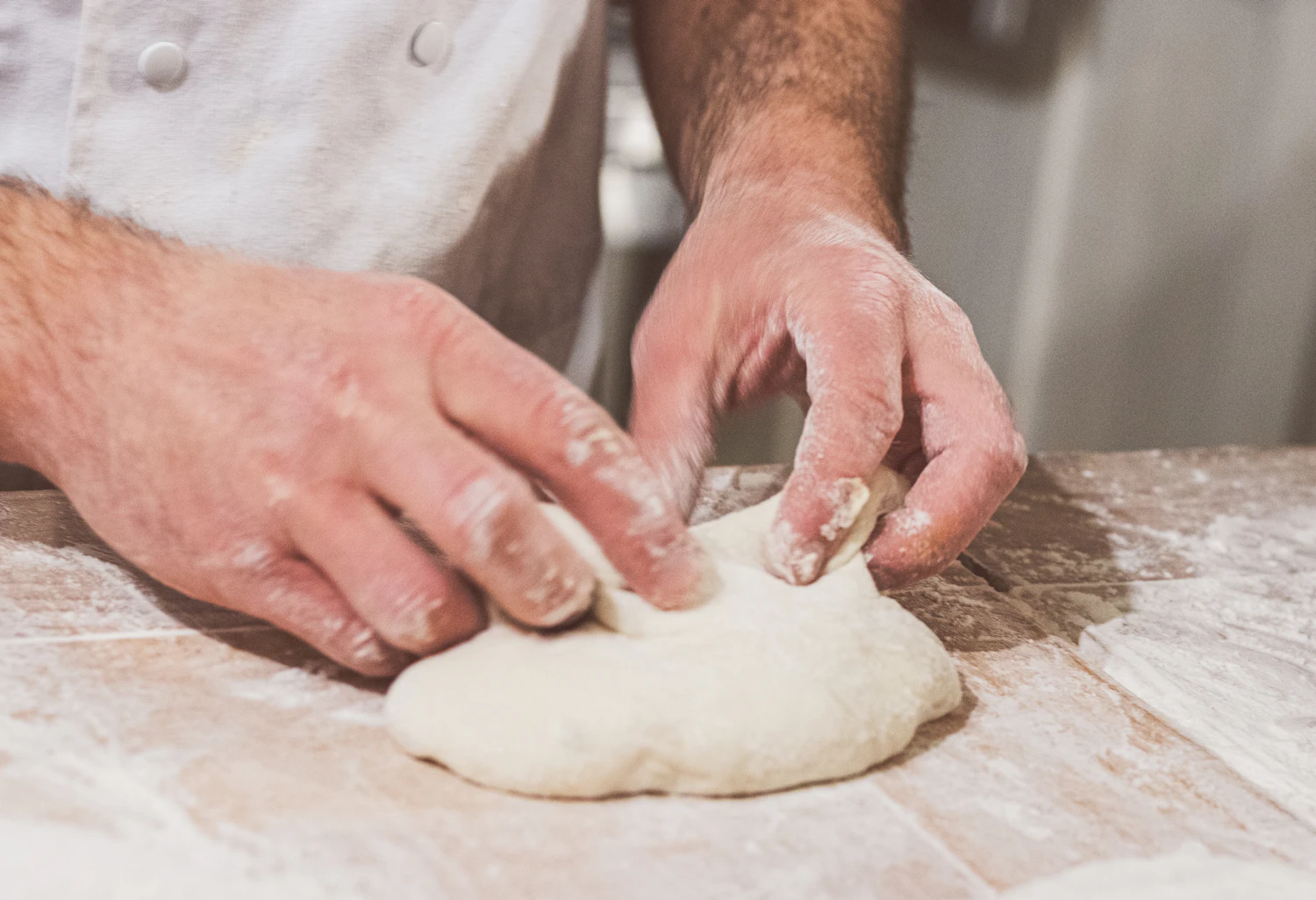 A baker's floured hands folding a pale round of dough on a wooden bench