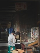 A vendor is seated on a green plastic stool, working at a street stall in a shaded area. The stall showcases an array of items, including lanyards and keychains, displayed on a table and hanging from a rack. The setting appears to be an indoor market or corridor with a partially closed metal gate behind the vendor. A sign overhead reads 'Koleksi Jaket Kulit' in Indonesian. The atmosphere is calm and casual, emphasizing everyday life.