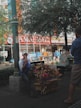 The image features a street scene with a vendor sitting beside a display of handmade toys, including wooden giraffes and other vibrant items. The vendor is wearing a cap and makes a gesture with his hand. In the background, a person wearing a mask is seated on a bench under a large tree. A building facade with various signs and storefronts is visible, suggesting an urban setting.