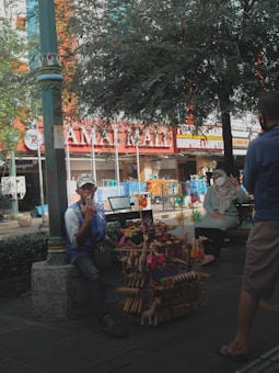 The image features a street scene with a vendor sitting beside a display of handmade toys, including wooden giraffes and other vibrant items. The vendor is wearing a cap and makes a gesture with his hand. In the background, a person wearing a mask is seated on a bench under a large tree. A building facade with various signs and storefronts is visible, suggesting an urban setting.