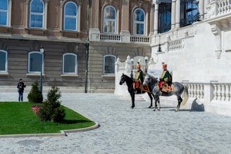 Two uniformed horsemen, dressed in historical military attire, are mounted on horses in front of an ornate, classical-style building with large windows and intricate stonework. The cobblestone path leads to a well-maintained lawn with small shrubs. A person stands to the side, taking photos or observing the scene.