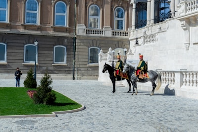 Two uniformed horsemen, dressed in historical military attire, are mounted on horses in front of an ornate, classical-style building with large windows and intricate stonework. The cobblestone path leads to a well-maintained lawn with small shrubs. A person stands to the side, taking photos or observing the scene.