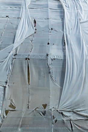 A building covered with a large, weathered tarp. The fabric is draped over scaffolding, showing signs of wear with holes and tears. The material hangs loosely, creating a textured and layered appearance.