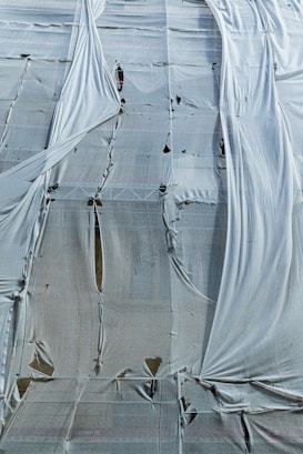 A building covered with a large, weathered tarp. The fabric is draped over scaffolding, showing signs of wear with holes and tears. The material hangs loosely, creating a textured and layered appearance.