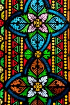 Vibrant stained glass design featuring symmetrical floral patterns in multiple colors. The intricate artwork includes interlocking geometric shapes and ornate details accentuated by bold outlines.