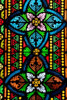Vibrant stained glass design featuring symmetrical floral patterns in multiple colors. The intricate artwork includes interlocking geometric shapes and ornate details accentuated by bold outlines.