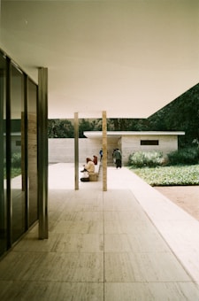 A minimalist architectural space with clean lines and large open areas. The interior blends with outdoor elements, featuring marble floors, columns, and glass walls. A few people are visible, some seated and others walking, suggesting a peaceful, contemplative environment. Lush greenery surrounds the area, adding a natural touch to the modern design.