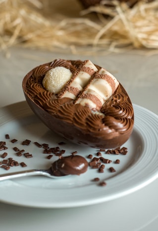 A beautifully crafted chocolate dessert resembling a half egg, filled with swirls of chocolate cream and topped with white chocolate decorations, placed on a white plate. Beside it is a spoon with chocolate and scattered pieces of chocolate.
