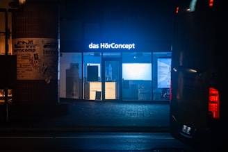 A storefront at night with bright blue lighting and a sign that reads 'das HörConcept.' The interior is illuminated, displaying various pieces of equipment or posters. Outside, a dark pillar to the left is covered with posters, and part of a vehicle is visible on the right side of the image.