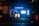 A storefront at night with bright blue lighting and a sign that reads 'das HörConcept.' The interior is illuminated, displaying various pieces of equipment or posters. Outside, a dark pillar to the left is covered with posters, and part of a vehicle is visible on the right side of the image.