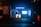A storefront at night with bright blue lighting and a sign that reads 'das HörConcept.' The interior is illuminated, displaying various pieces of equipment or posters. Outside, a dark pillar to the left is covered with posters, and part of a vehicle is visible on the right side of the image.