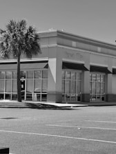 A monochrome photograph of a commercial building with large windows and awnings. In the foreground, there is a solitary palm tree, and the parking area appears empty.