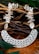 An intricately designed necklace featuring a series of white shell-like beads connected to a large, textured pendant with geometric cutouts. The background consists of interwoven natural wood sticks and green leaves, adding an organic and earthy feel to the image.