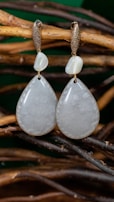 A pair of elegant teardrop-shaped earrings hanging on intertwined natural wooden branches. The earrings have a smooth, lustrous appearance with a subtly shimmering texture. Above each teardrop is a smaller, irregularly shaped bead, and both hang from gold-colored hooks with a rough, textured finish.